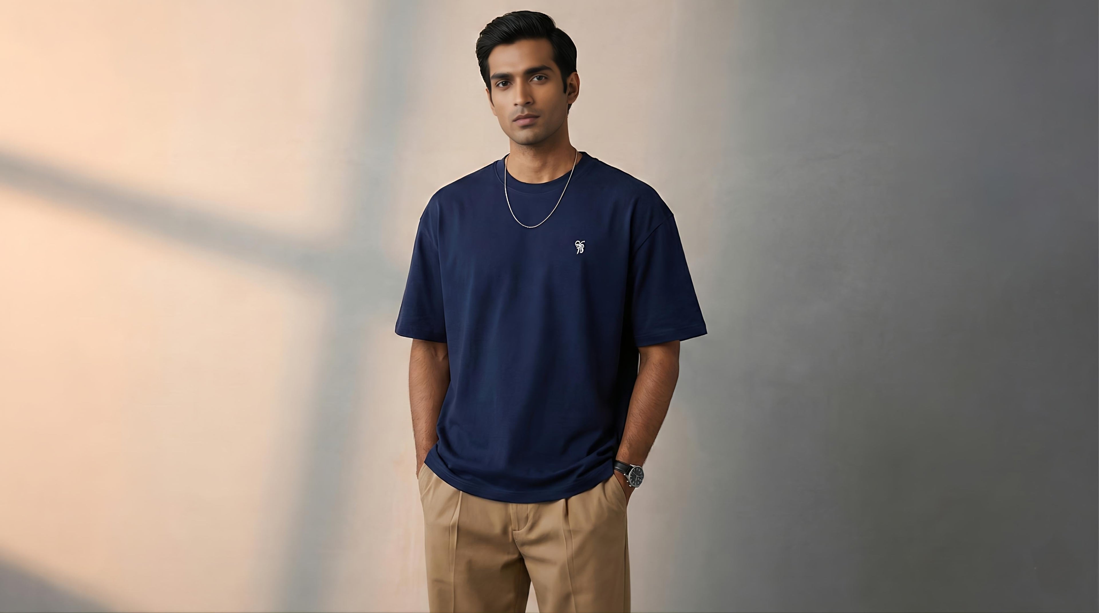 A male model standing confidently in a studio environment wearing a premium navy blue oversized T-shirt and tan trousers under soft, warm lighting.