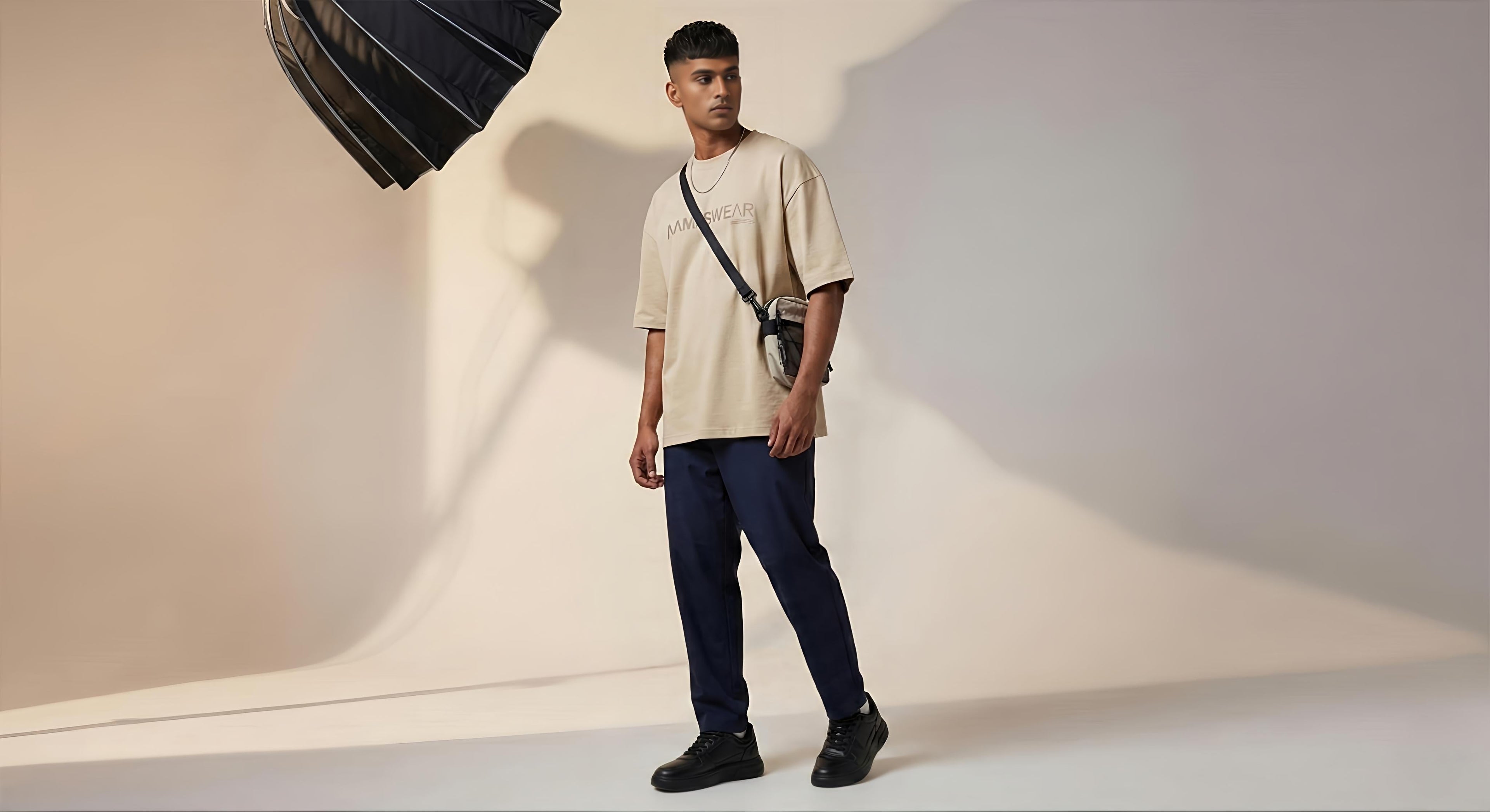 Full-body studio shot of a male model wearing an oversized beige t-shirt with "AAMS WEAR" branding on the chest, paired with navy blue pants and black chunky sneakers. A photography softbox is visible in the upper corner.