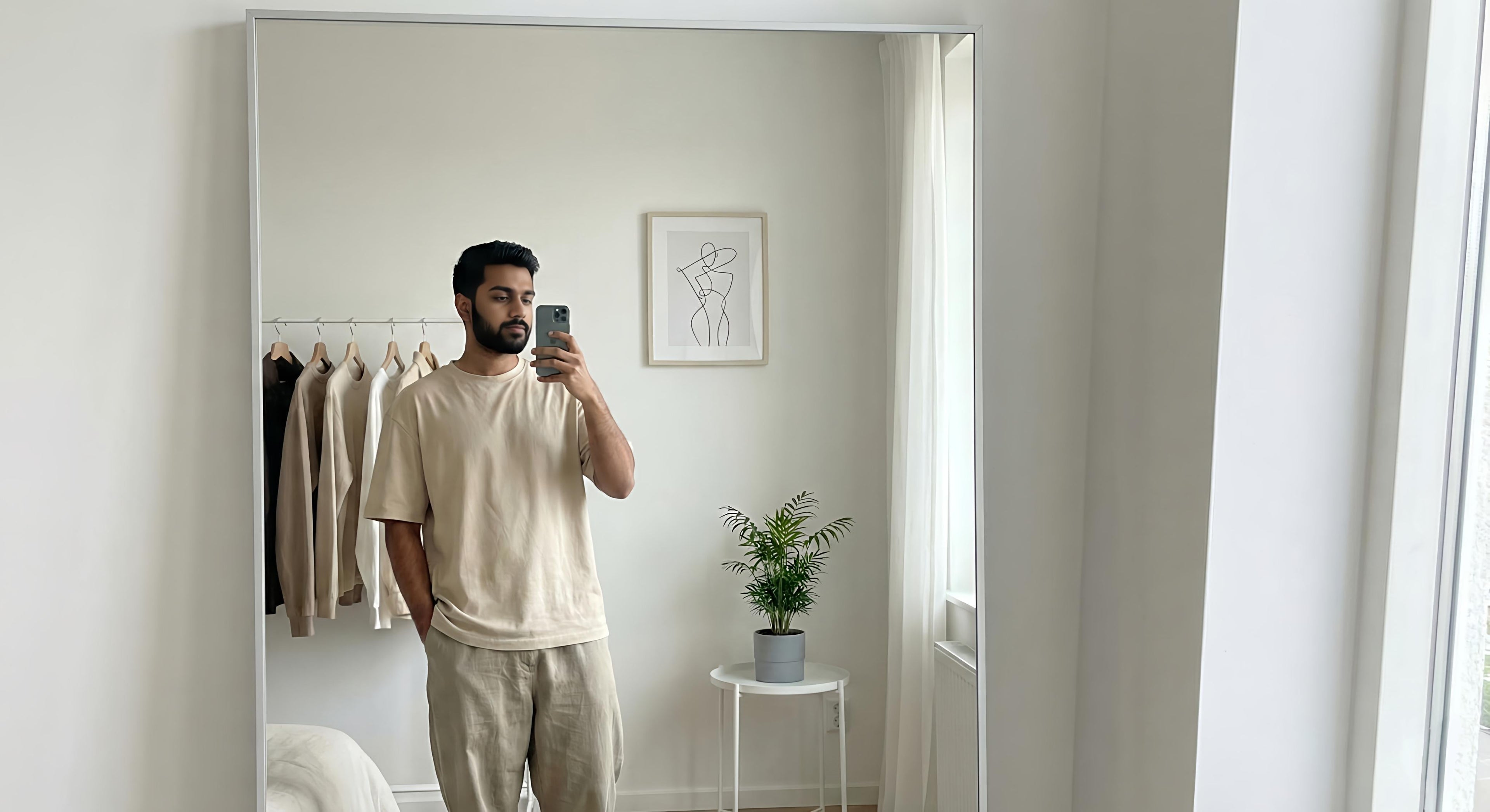 Indian fashion influencer taking a mirror selfie in a minimal bedroom, wearing a clean beige oversized T-shirt and loose trousers, with a clothing rack and modern decor in the background.
