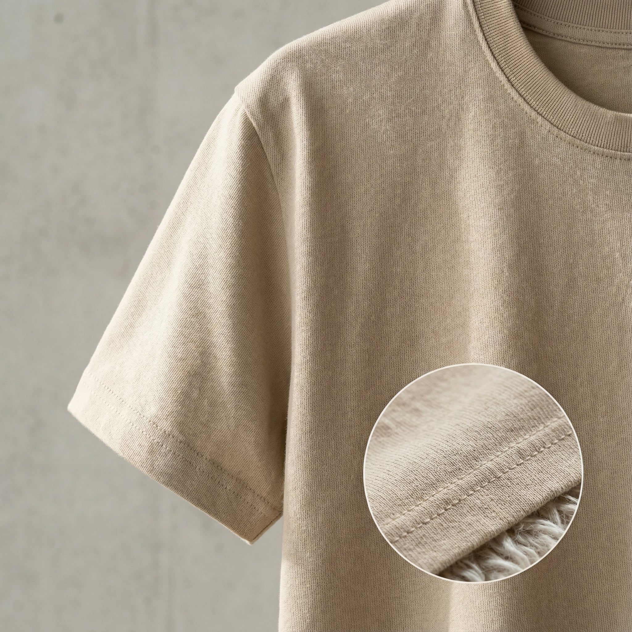 A close-up photograph of a beige, crew neck t-shirt hanging against a grey concrete wall, focusing on the sleeve.