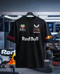 Full front view of an oversized black Red Bull Racing T-shirt hanging on a hanger against a blurred F1 garage background. The shirt features sponsor logos on the chest, large "Red Bull" text across the center, "RB21" in red outline on the right sleeve, and the charging bulls logo on the left sleeve.