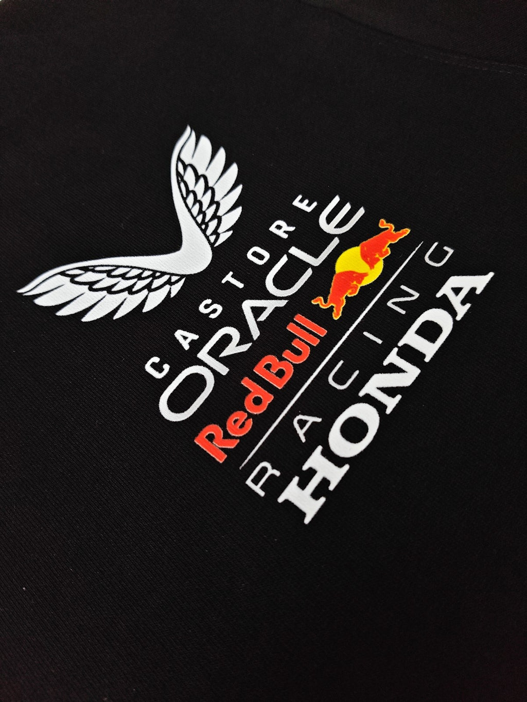 Macro detail shot of the chest logo stack on black fabric, featuring the winged Castore logo above "Oracle Red Bull Racing Honda" text in white, with the iconic red and yellow bulls logo.