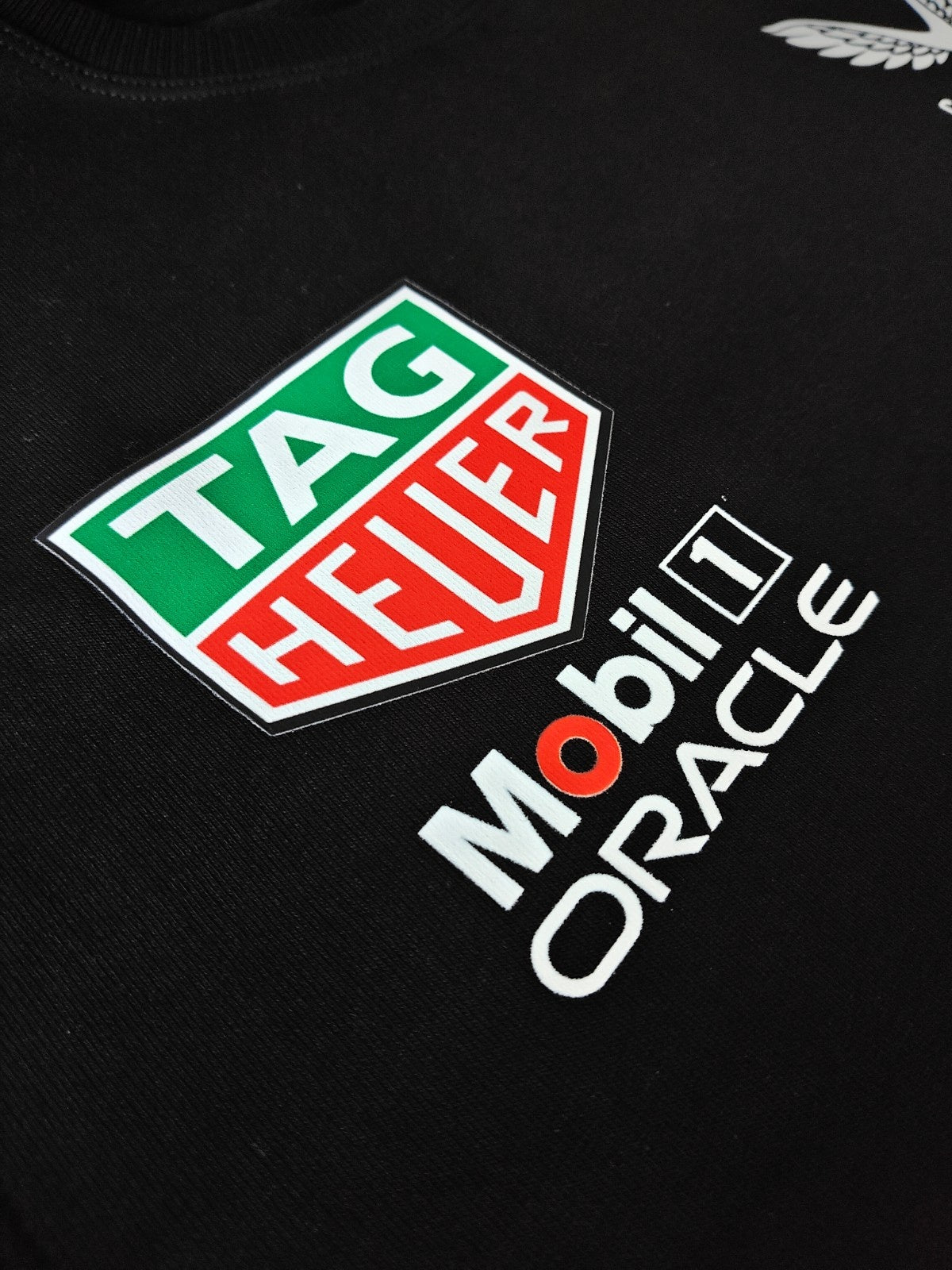 Close-up of the right chest area showing the green and red TAG Heuer shield logo, stacked above white Mobil 1 and Oracle sponsor text on a black T-shirt.