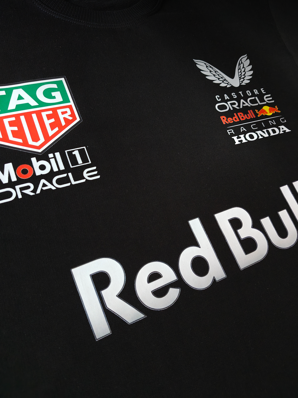 Close-up view of the upper chest on a black Red Bull Racing T-shirt. The image displays the Castore, Oracle, Red Bull Racing, and Honda logos on the left chest, and the TAG Heuer, Mobil 1, and Oracle sponsor logos on the right chest, sitting above large silver "Red Bull" lettering.