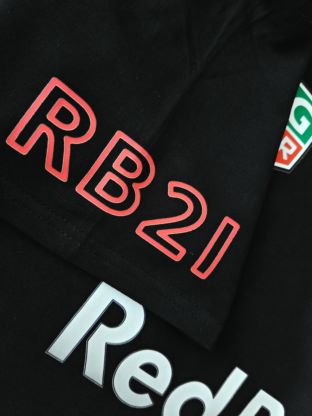 Detail shot of the T-shirt sleeve featuring "RB21" text printed in a red outline font on black cotton fabric, with a partial view of the silver central chest text below.