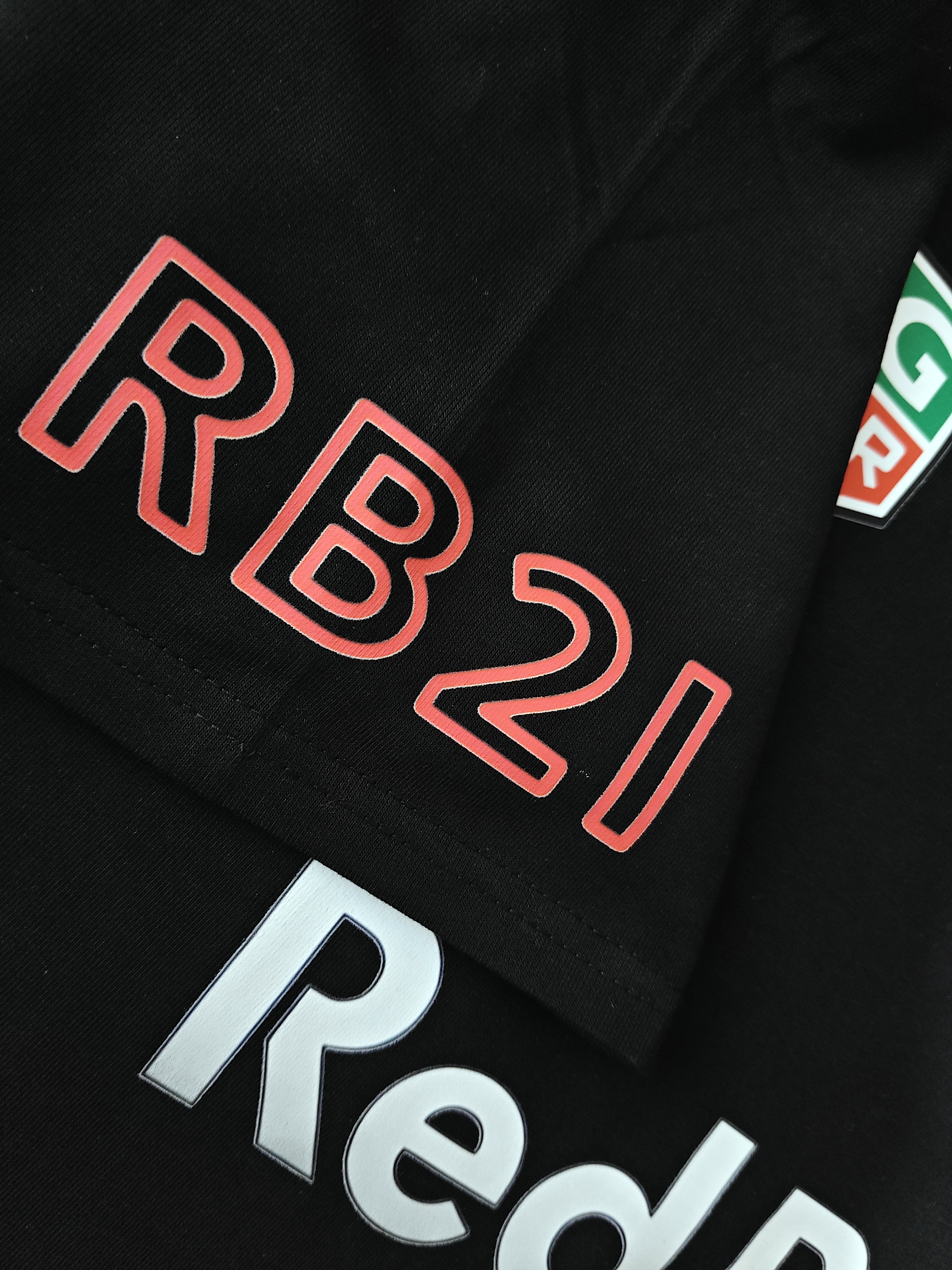 Detail shot of the T-shirt sleeve featuring "RB21" text printed in a red outline font on black cotton fabric, with a partial view of the silver central chest text below.